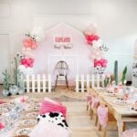 Pink barn backdrop with balloons, cow-print accents and a white picket fence at Lyla’s first rodeo birthday party