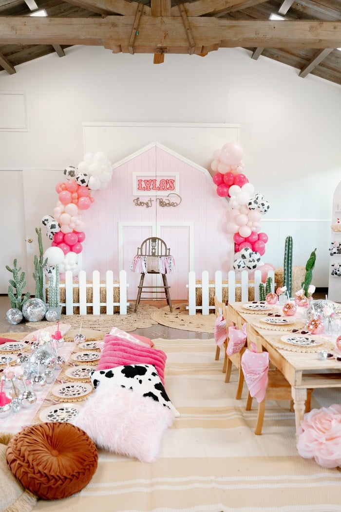 Pink barn backdrop with balloons, cow-print accents and a white picket fence at Lyla’s first rodeo birthday party